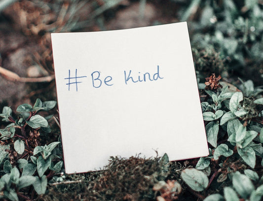 words #be kind on a backdrop of little plants on the forest floor